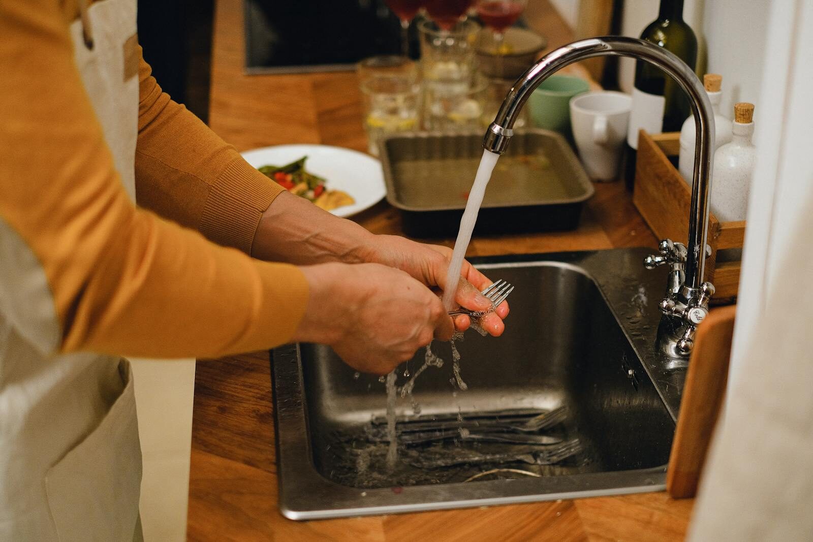Close-up of hands washing a fork under running water in a kitchen sink with a cozy countertop setup.
