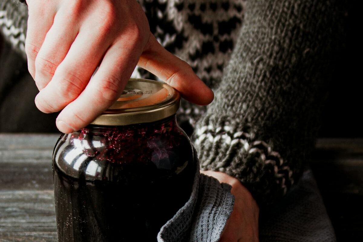 Close-up of hands opening a glass jar on a rustic table, showcasing homemade preserves in cozy kitchen light.