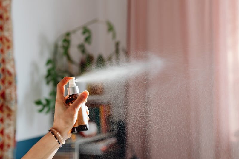 A woman's hand spraying fragrance in a cozy domestic room with elegant curtains.